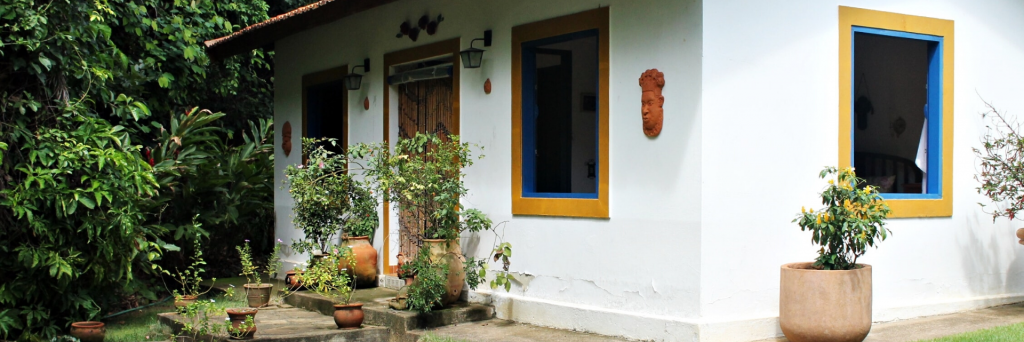 A picture of our gite: a small white house with large blue windows surrounded by plants and hedges.